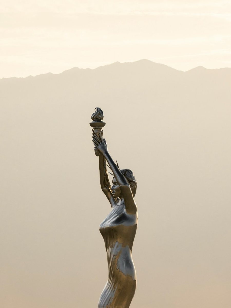 A tall statue holding up a torch in a misty desert at sunrise