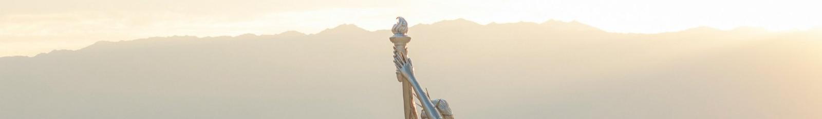 A tall silver statue raising a torch in a hazy desert at sunrise